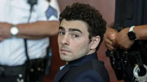 Luigi Mangione, dressed in a dark suit and blue chequered shirt, looks at the camera with law enforcement officials in the background