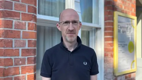 A bald man wearing glasses and a navy blue polo shirt. He is standing outside a railway station. 