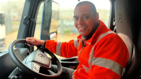 Fiona Irving / BBC A man with a shaved head, wearing a hi-vis orange jacket smiles at the camera. He is sitting in the driving seat of a HGV cab and has one hand on the steering-wheel.
