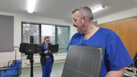 A mobile X-ray kit has been set up in a large grey room. A man and a woman are in the room wearing dark blue medical scrubs. The man, in the foreground, who has short grey hair, a beard, and glasses, is holding an X-ray imaging panel. In the background, a woman with long hair tied back is working on a laptop. An imaging device is mounted on a tripod in the room.