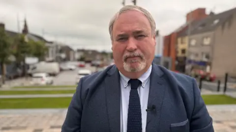 Colm Gildernew is standing outside. A road is behind out of focus. He's wearing a navy suit and tie and a blue shirt. 