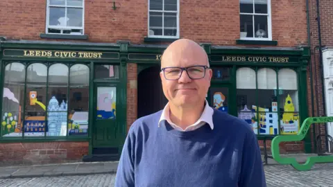BBC/Sofía Luis-Hobbs A man in a blue jumper and blue glasses smiles at the camera as he stands in front of the Leeds Civic Trust building. The unit has a green front, with colourful drawings of the city in its windows, and Leeds Civic Trust painted on in gold writing above.