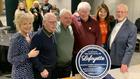 Wolverhampton Society Six people gathered around a blue plaque. They are at an event and there are people standing around drinking prosecco. The plaque says "Lafayette" and has the branding of the Wolverhampton Society.
