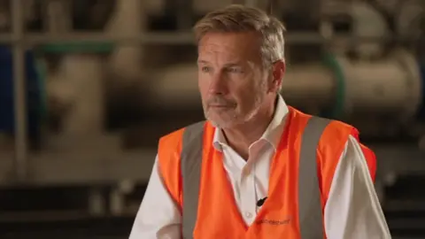 BBC A man with greying short brown hair and stubble, wearing a white shirt and orange high visibility vest. It has South East Water written on it. 