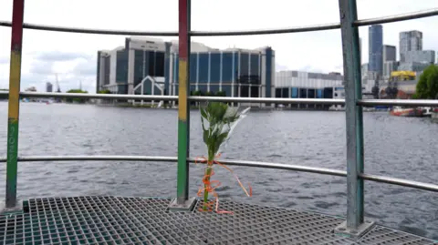 Bouquet of flowers tied to a metal railing overlooking a river, with modern buildings in the background.