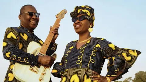 Ojoz Music act Amadou and Mariam smiling. One man and one woman both in black and yellow outfits. 