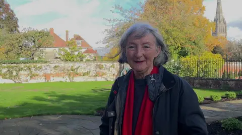 Liz Slater, from Salisbury Cathedral Close Preservation Society, stands in the garden of a neighbouring property to Leaden Hall. Behind her right shoulder can be seen the terracotta coloured roof of the Georgian house and over her left shoulder the spire of Salisbury Cathedral. The brick wall surrounding Leaden Hall can be seen adjoining the lawn of the garden.