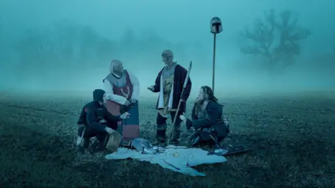 Simon Plant Four people dressed in medieval clothes are sitting on a field. It is dark and fog is coming up from the ground.