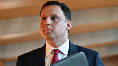 Anas Sarwar, who has short black hair, looks to his right in a close-up shot. He is wearing a black suit, white shirt and red tie, and holding a folder to his chest. There is wood panelling out of focus behind him. 