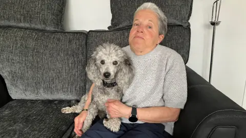 An woman sits on a dark grey sofa holding a small curly-haired dog on her lap. The dog looks directly at the camera while wearing a collar, with soft textured fur visible.