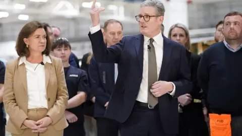 Reuters Eluned Morgan and Keir Starmer on a visit, standing in front of site workers. Eluned is on the left in a beige suit with a white shirt and Keir is on the right with his right hand in the air, wearing a blue suit, white shirt and brown tie. He has glasses on.