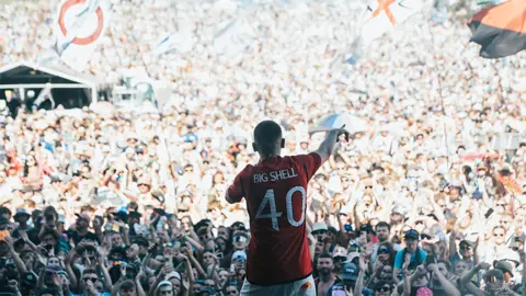 Adidas Aitch wearing the new Manchester United shirt, which is red. The back of the shirt says Big Shell with the number 40 below it. He is wearing white trousers, and is holding a microphone, with his right arm pointing at the crowd in front of him, full of thousands of people watching him perform. There are big flags in the crowd and many people on their phones filming his performance.