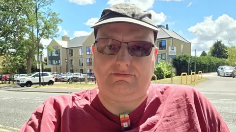 Doug Paulley A close-up outdoor selfie of a man wearing a green cap, rectangular tinted glasses, and a maroon T-shirt. He is centered in the frame with a neutral expression, on a sunny street with parked cars, apartment buildings, trees, and a bright blue sky with clouds in the background.