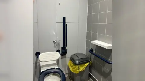 TransPennine Express A toilet, a bin and a shelf in a railway station bathroom. The walls are white and grey.