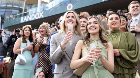 PA Media Two women, one in a grey double-breasted trouser suit and one in a green halter-necked dress, hold glasses and stand at the front of a crowded stand at Aintree