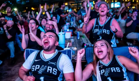 Brandon Bell/Getty Images People sitting outdoors face the camera wearing Spurs jerseys looking animated and smiling while cheering