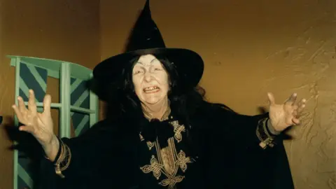 Ben Willis A woman wearing a black witch's hat and black cloak, snarls at the camera with her hands outstretched.