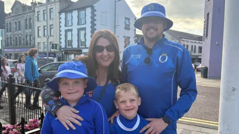 Jude Johnston, Mathew Williams and family out celebrating the Dungannon Swifts. Jude has long brown hair and wearing sunglasses and blue top. Matthew is wearing a blue Swifts half zip and hat. Their two boys are standing with them.