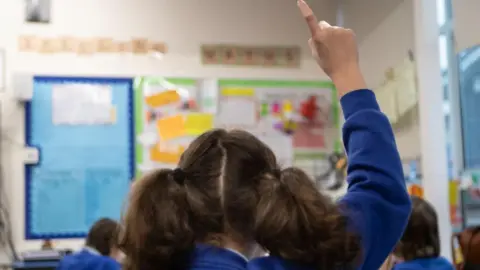 PA Media A schoolgirl, photographed from behind, raising her hand in class. The girl is in year 5 and has brown hair in bunches and is wearing a royal blue cardigan. The backs of other children's heads, also wearing royal blue jumpers are cardigans, can be seen as they sit at tables. A colourful display can be seen out focus on the far wall.