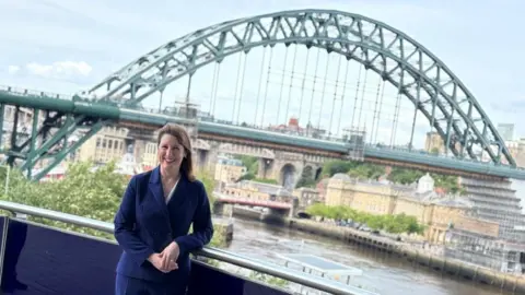 LDRS Rachel Reeves, who has dark brown hair, is wearing a navy blue suit and is standing on a balcony, leaning against a railing. She is smiling and facing the camera. Behind her is the green Tyne Bridge. It is almost fully visible with part of both the High Level Bridge and Swing Bridge in view behind it. Reeves stands on the Gateshead side of the river and part of Newcastle Quayside can also be seen.