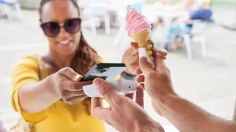 Una mujer que usa gafas de sol con gafas de sol con un helado rosa colocando su teléfono inteligente en la terminal de pago.