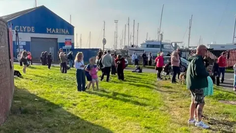 A crowd of people stands on a grassy area beside Clyde Marina, with yachts and sailboat masts visible behind a fence in the background. A large blue building marked “CLYDE MARINA” stands to the left, while people gather along the roadside and waterfront under bright sunshine. A speed limit sign and several parked vehicles are visible near the marina entrance.