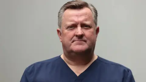 PA Media A man looks into the camera with a straight face. He has brown and white hair combed to the side. He is wearing dark blue medical scrubs.