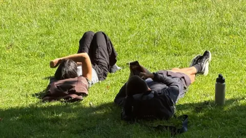 Two people lying down in the grass. The girl on the left is lying in the sun while the man to the right is partially shaded by a tree.