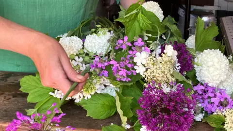 A close up of a pair of hands working on a flower arrangement. The arrangement contains pink, purple and white flowers with green leaves.