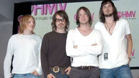 Getty Images The set  Kasabian. All 4  members are dressed due  to the era, 2004, which includes choky  achromatic  low-cut t-shirts, wavy haircuts and crisp  facial hair.