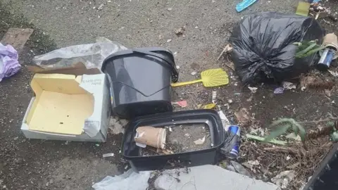 Supplied Rubbish including a bin bag, a box, paper cups, a mop bucket, a fly swatter and drinks cans is scattered across a pavement. 
