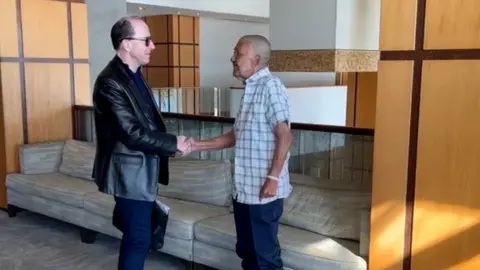 Matthew Gayle Mark, wearing a leather jacket and dark sunglass shakes hands with Hosein, who is wearing a checked shirt and jeans