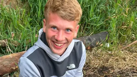 Lucas Littlefair, a teenager who died in a crash in Hesleden. He has blonde hair and blue eyes and he is looking up at the camera with a wide smile. He is wearing a grey The North Face hoodie and is in a green area, with a tree trunk on the ground behind him and some grass.