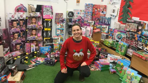 Jennha Fansa, a woman with log dark hair tied back, wears a red jumper with a Christmas pudding design. She crouches in front of a display of toys in storage, smiling.