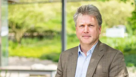 Jeff Dauling/Embl Ebie, Professor-Men, who has greyish brown hair, looks at the camera, putting a blue shirt and a brown suit-lace blurred green background trees and shrubs for HIME