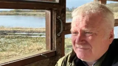 Guy Campbell/BBC A person wearing a green jacket sits in a bird hide staring out over a marshland scene