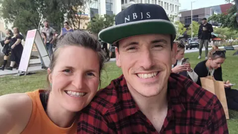 Jess Gray and Matt Kidd -the couple are sitting together and smiling. Jess has an orange top on and dark hair, Matt has a red and black chequed shirt and a black baseball cap on. They are sitting outside.  