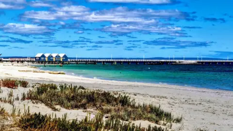 Getty Images A bright blue beach is pictured with four huts along a pier. It's a sunny day. 