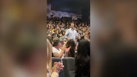 Man in a light blue shirt walking down the steps of an auditorium as the audience applause.
