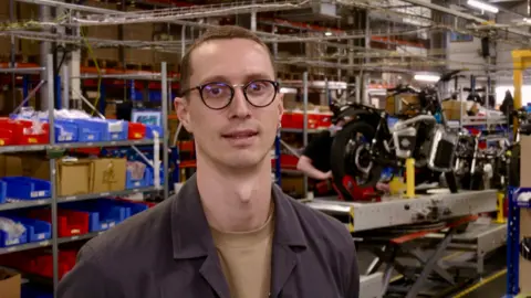 A man with short dark hair and wearing glasses is looking at the camera. Boxes are on shelves behind him on the left of the photo.