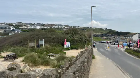 Mawgan Porth with picnic benches to the left of the foreground, a road, people and shops to the right and houses in the background