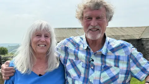 A man and a woman stand close together, smiling at the camera, with his arm around her shoulders. The woman has long white hair and is wearing a blue t-shirt; the man has greying curly hair and a grey beard and is wearing a blue and white checked shirt. A low farm building and pale sky fill the background.
