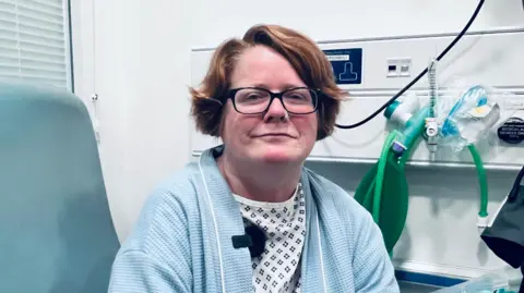Patient Lisa Brittain sits in a chair on a ward. She is wearing a hospital gown and dressing gown. She wears glasses and is smiling at the camera.