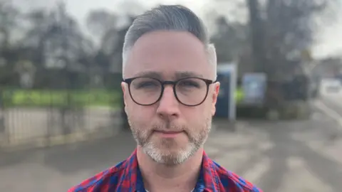 A man looks directly at the camera. He is wearing glasses and a red plaids shirt. He has grey hair and light stubble. 