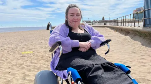 A woman with brown hair is sitting in a blue beach wheelchair, with a sandy beach, the sea and a promenade seen behind her. She is wearing a purple cardigan and a black dress.