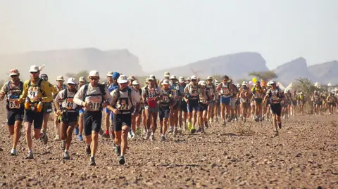 Getty Images A large group of runners race across the Sahara Desert in bright sunshine.