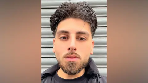 Ismail Qureshi has black medium-length hair and is looking straight at the camera, he has a short brown/black beard and a black T-shirt with a black jacket over the top. He is standing in front of a metal shutter.