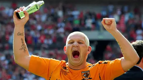 Reuters Dean Windass with arms raised aloft on the pitch at the Wembley play-off final. He holds a trophy in his right hand and is wearing his amber Hull City shirt