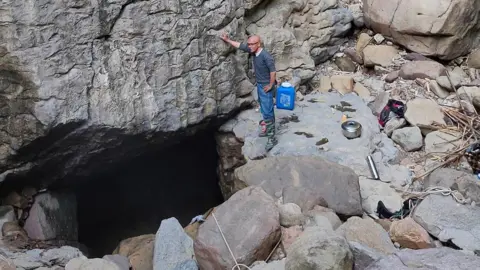 Khlur Baiaineh Mukhim A man stands next to a cavernous hole surrounded by boulders, on a hill in Meghalaya.