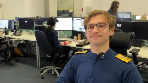 A man wearing a navy blue top with yellow shoulder pads. He is sat in an office with people and computers in the background.